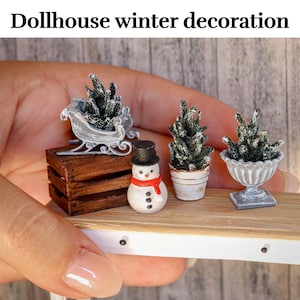 Könnte beinhalten: Eine Miniatur-Winterlandschaft mit einem Schneemann, einer Holzkiste und drei kleinen Topfpflanzen. Die Bäume sind mit weißem Flock bedeckt, um Schnee zu imitieren. Die Szene ist auf einem Holzregal platziert.