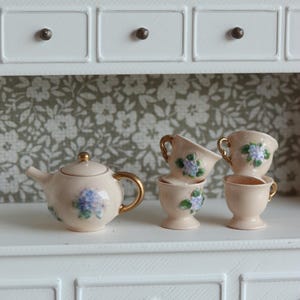 Könnte beinhalten: Ein miniaturisiertes Teeservice in Hellrosa mit Teekanne und Tassen. Das Set hat goldfarbene Akzente und blaue Blumenmuster. Es steht auf einem weißen Regal vor einer Tapete mit floralem Muster.