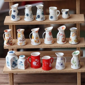 Può includere: Una mensola di legno espone una collezione di brocche e vasetti in ceramica in miniatura. Le brocche presentano disegni floreali, motivi autunnali e accenti rossi. La collezione comprende varie dimensioni e forme, mostrando una varietà di stili decorativi.