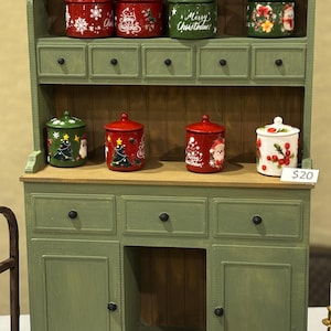 Può includere: Una credenza in legno verde salvia con diversi cassetti e ripiani. I ripiani ospitano otto barattoli con coperchio decorati a tema natalizio con la scritta Merry Christmas. Quattro barattoli aggiuntivi e una brocca sono davanti.