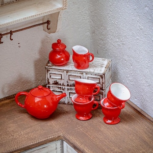Könnte beinhalten: Ein Miniatur-Teeservice aus roter Keramik, bestehend aus einer Teekanne mit Deckel, einer Zuckerdose mit Deckel, einem Milchkännchen und vier Teetassen auf Standfuß. Das Set hat eine glänzend rote Außenfläche und eine weiße Innenglasur.
