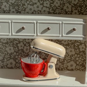 Può includere: Un mixer a immersione in miniatura color crema con una frusta argentata e una ciotola rossa brillante. L'elettrodomestico presenta una finitura liscia e lucida e poggia su una base, progettato come elemento decorativo per una cucina di bambole.