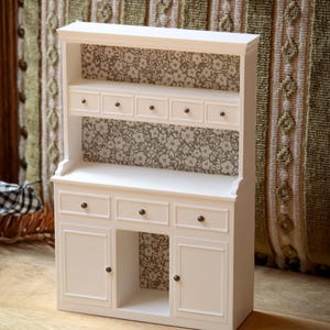 Può includere: Una credenza in miniatura bianca con un ripiano superiore dotato di cinque piccoli cassetti e una sezione inferiore con tre cassetti e due ante. I pannelli posteriori presentano un motivo floreale marrone e bianco, con pomelli su ogni anta.