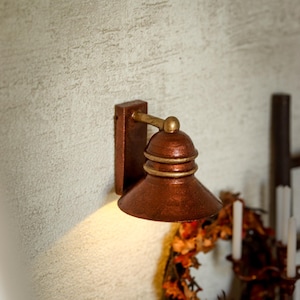Könnte beinhalten: Eine Wandleuchte mit metallisch kupferfarbenem Finish und glockenförmigem Schirm. Die Leuchte verfügt über eine rechteckige Grundplatte, einen kurzen horizontalen Arm und zwei goldfarbene Ringe, die den kuppelförmigen Schirm verzieren.