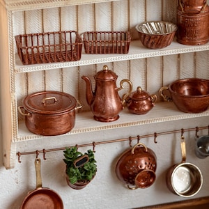 May include: Miniature copper kitchenware set displayed on a white shelf. Includes pots, pans, a teapot, and a colander. The set also features small baskets and a Farmers Market container. A charming, detailed display.