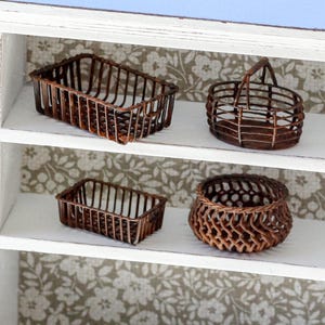 Puede incluir: Cuatro cestas miniatura tejidas de color marrón dispuestas en dos estantes blancos. El estante superior contiene una cesta rectangular y una cesta redonda con asa. El estante inferior presenta una cesta rectangular más pequeña y una cesta redonda.