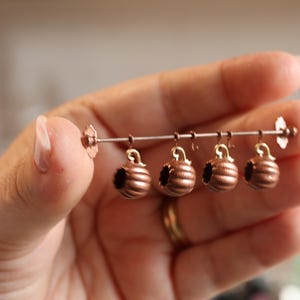 Può includere: Una barra di metallo color rame fatta a mano con quattro ornamenti miniaturizzati a forma di campana, a coste, appesi. Gli ornamenti hanno un piccolo anello nella parte superiore. La barra ha accenti floreali decorativi su ogni estremità.