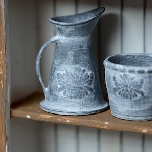 Könnte beinhalten: Ein grauer Metallkrug und ein passender zylindrischer Topf, beide mit einem abgenutzten Finish und einem erhabenen Blumenrelief auf der Vorderseite. Der Krug hat einen geschwungenen Griff und einen Ausguss, während der Topf eine offene Form hat.