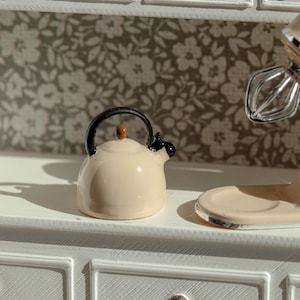 Könnte beinhalten: Eine cremefarbene Miniatur-Teekanne aus Keramik mit glänzender Oberfläche, schwarzem Griff und schwarzem Ausguss. Die Teekanne hat einen kleinen braunen Knopf auf dem Deckel und steht auf einer weißen Oberfläche neben einem Teil eines Standmixers.