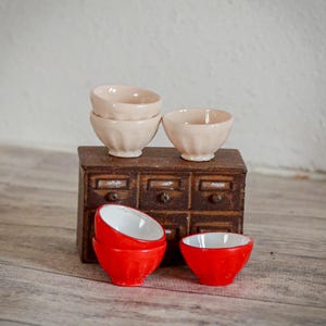 Pode incluir: Um conjunto de taças de cerâmica em miniatura nas cores creme e vermelho brilhante, com um design exterior canelado e interiores brancos lisos, dispostas sobre e em frente a um pequeno armário de madeira castanho-escuro com gavetas.