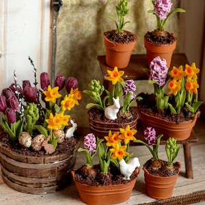 Op de afbeelding: Een verzameling miniatuurplanten in potten met paarse tulpen, gele narcissen en roze hyacinten in terracotta en houten bakjes. Het arrangement bevat kleine witte keramische dierenfiguurtjes en gespikkelde eieren in de aarde.