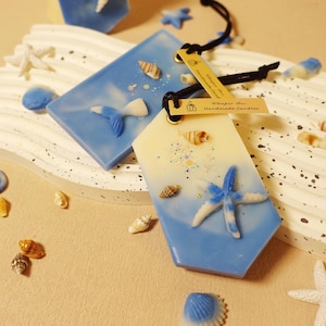 Pode incluir: Duas velas artesanais com tema de praia. Uma é um gradiente azul e branco com uma estrela do mar, a outra é azul com uma cauda de sereia. Pequenas conchas e estrelas do mar estão espalhadas.