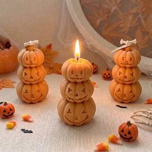 May include: Three stacked orange pumpkin candles with carved jack-o'-lantern faces. The centre candle is lit with a bright flame. The candles are surrounded by small decorative pumpkins, candy corn, a skeleton hand, and black bat silhouettes.