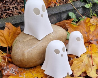 Könnte beinhalten: Drei weiße Geisterfiguren mit ausgeschnittenen Augen und Mündern werden auf einem Stein ausgestellt und von Herbstblättern umgeben. Die Geister sind unterschiedlich groß, wobei der größte auf dem Felsen sitzt.