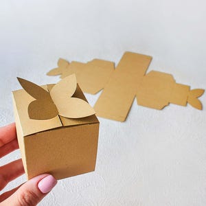 Op de afbeelding: Een kleine, geassembleerde bruine kartonnen geschenkdoos met vlindervormige uitsparingen aan de bovenkant. De doos wordt vastgehouden door een hand met roze nagellak. Uitgevouwen kartonnen stukken zijn op de achtergrond.