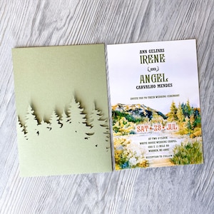 Könnte beinhalten: Hochzeitseinladung mit einer grünen Waldsilhouette auf der Vorderseite und einer Aquarelllandschaft mit dem Text "ANN GELINAS IRENE UND ANGEL CARVALHO MENDES" auf der Rückseite. Die Einladung lautet "INVITE YOU TO THEIR WEDDING CEREMONY SAT 28 JUL AT TWO O'CLOCK WHITE HOUSE WEDDING CHAPEL 4360 E 13 MILE RD WARREN, MI 48092 RECEPTION TO FOLLOW."
