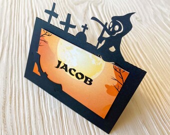 Könnte beinhalten: Schwarze Papier-Platzkarte mit einem gruseligen Halloween-Motiv. Die Karte zeigt eine Silhouette eines Sensenmanns mit einer Sense, einen Mond und drei Kreuze. Der Name "Jacob" ist auf die Karte geschrieben.
