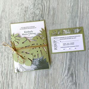 Pode incluir: Convite de casamento verde cortado a laser com um design de folhas tropicais. O convite inclui os nomes Kimberly Marie Uftring e Christophe. O cartão RSVP solicita uma resposta até 15 de maio de 2019.