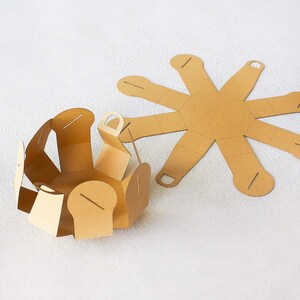 May include: A brown cardboard box with a starburst shape that folds into a round container. The box is flat and unfolded in the background, and folded into a round shape in the foreground.