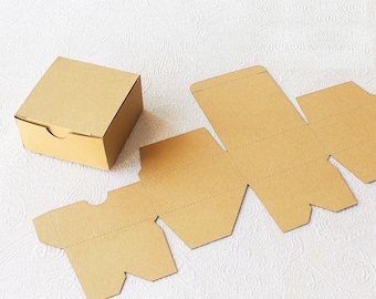 May include: A square, assembled cardboard box with a lid, next to the unfolded, die-cut pieces of a similar box. The boxes are a light brown color and are shown on a white patterned surface. The box is designed for packaging and shipping.