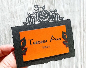 Könnte beinhalten: Schwarze und orangefarbene Halloween-Platzkarte mit einem ausgeschnittenen Design aus Kürbissen und Blättern. Die Karte lautet "Theresa Ann, Tisch 5".