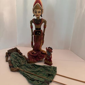 May include: A collection of Indonesian Wayang Golek puppets. The puppets are hand-carved and painted with intricate details. The main puppet is a standing figure with a red and gold headdress and a red and gold skirt. The other puppets are lying down and standing.