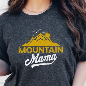 May include: Dark gray heather t-shirt with a yellow mountain graphic and the words "MOUNTAIN Mama" in white script. The shirt is worn with blue jeans. Additional text includes "Fast shipping" and "Next Day Shipping".