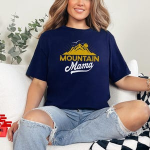 May include: Navy blue t-shirt with a gold mountain graphic and the text "MOUNTAIN Mama" in white script. The shirt is a relaxed fit and is available in sizes 3XL-5XL.