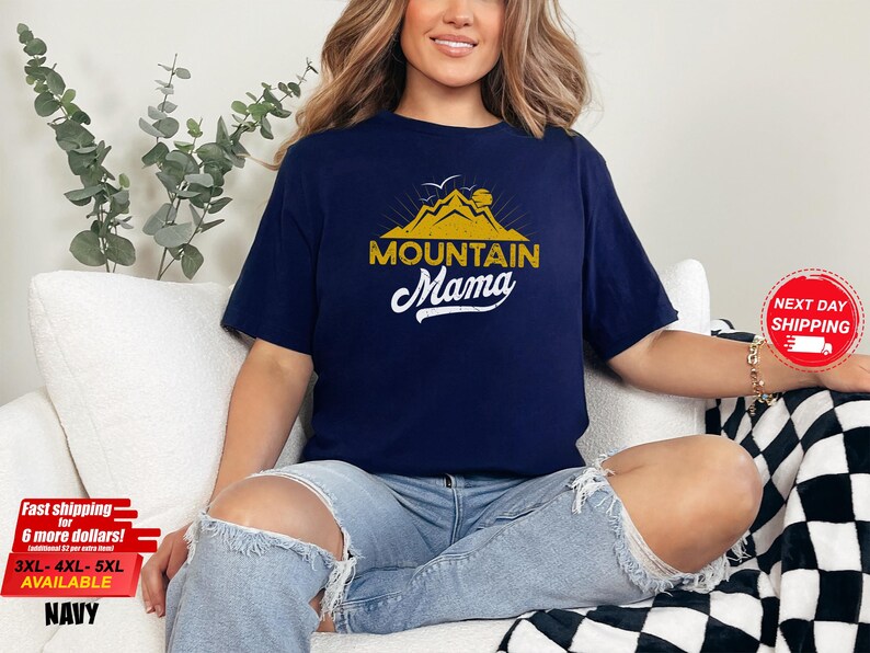 May include: Navy blue t-shirt with a gold mountain graphic and the text "MOUNTAIN Mama" in white script. The shirt is a relaxed fit and is available in sizes 3XL-5XL.