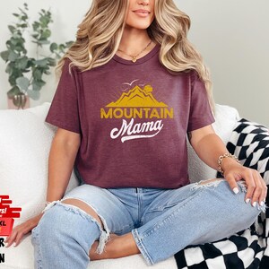 May include: Maroon heather t-shirt with the text "Mountain Mama" in gold and white. The shirt features a mountain and sun graphic. Available in sizes 3XL, 4XL, and 5XL. Fast shipping is available.