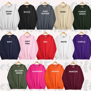 May include: A collection of crewneck sweatshirts in various colors, including sport gray, black, dark heather, sand, forest green, navy, soft pink, red, white, purple, military green, raspberry, orange, and maroon. Each sweatshirt is displayed on a wooden hanger.