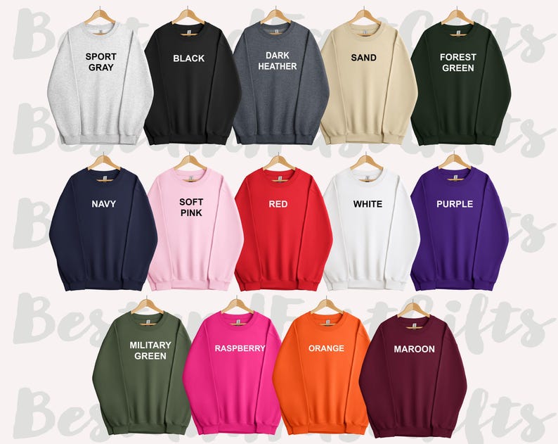 May include: A collection of crewneck sweatshirts in various colors, including sport gray, black, dark heather, sand, forest green, navy, soft pink, red, white, purple, military green, raspberry, orange, and maroon. Each sweatshirt is displayed on a wooden hanger.