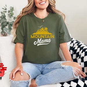 May include: Olive green t-shirt with the text "MOUNTAIN Mama" in a yellow and white design. The shirt features a mountain graphic. The shirt is available in sizes 3XL, 4XL, and 5XL.