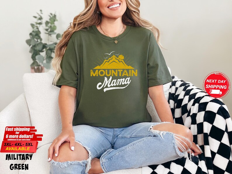 May include: Olive green t-shirt with the text "MOUNTAIN Mama" in a yellow and white design. The shirt features a mountain graphic. The shirt is available in sizes 3XL, 4XL, and 5XL.