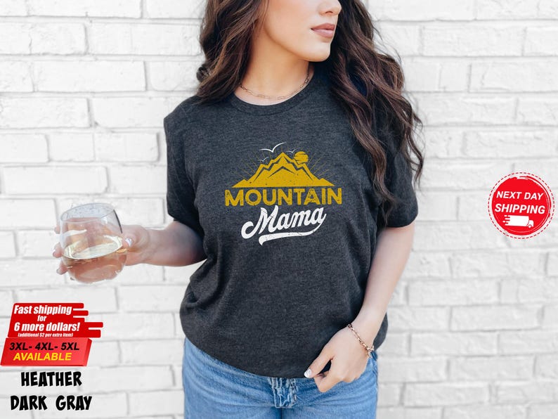 May include: Dark gray heather t-shirt with a yellow mountain graphic and the words "MOUNTAIN Mama" in white script. The shirt is worn with blue jeans. Additional text includes "Fast shipping" and "Next Day Shipping".