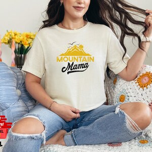 May include: Cream-colored t-shirt with the text "Mountain Mama" in black script and yellow block letters, featuring a mountain graphic. The shirt is worn with blue jeans. The image also includes a floral arrangement and a decorative pillow.