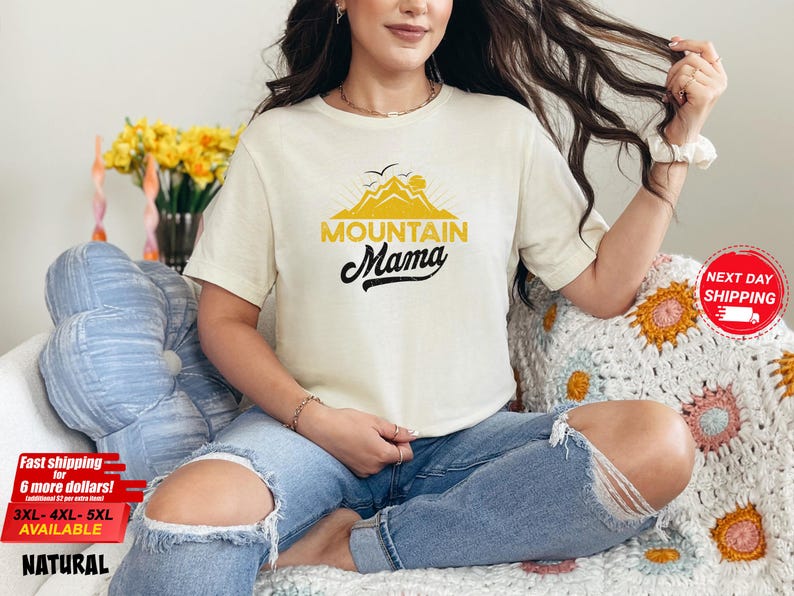 May include: Cream-colored t-shirt with the text "Mountain Mama" in black script and yellow block letters, featuring a mountain graphic. The shirt is worn with blue jeans. The image also includes a floral arrangement and a decorative pillow.