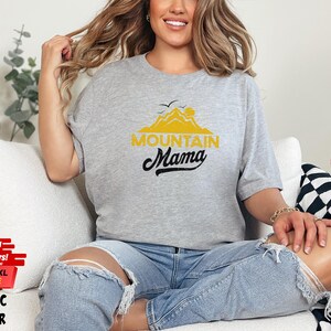 May include: A light gray athletic heather t-shirt with the text "MOUNTAIN MAMA" in yellow and black. The shirt features a mountain and sun graphic. The model is wearing ripped blue jeans.