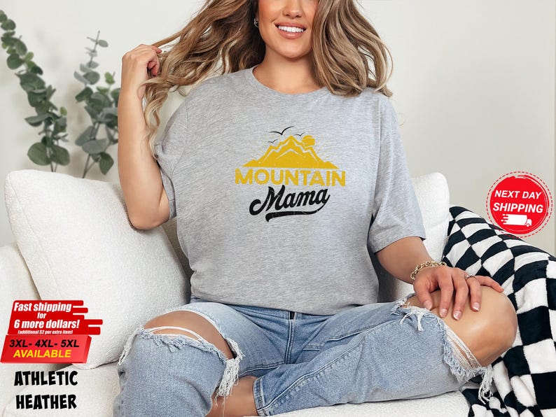 May include: A light gray athletic heather t-shirt with the text "MOUNTAIN MAMA" in yellow and black. The shirt features a mountain and sun graphic. The model is wearing ripped blue jeans.