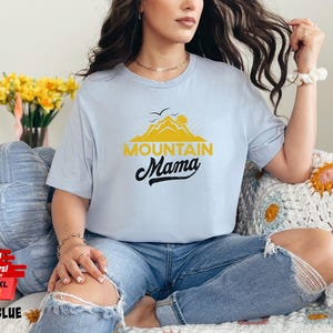 May include: Light blue t-shirt with the text "Mountain Mama" in black script and a yellow mountain graphic. The shirt is light blue and is available in sizes 3XL, 4XL, and 5XL.