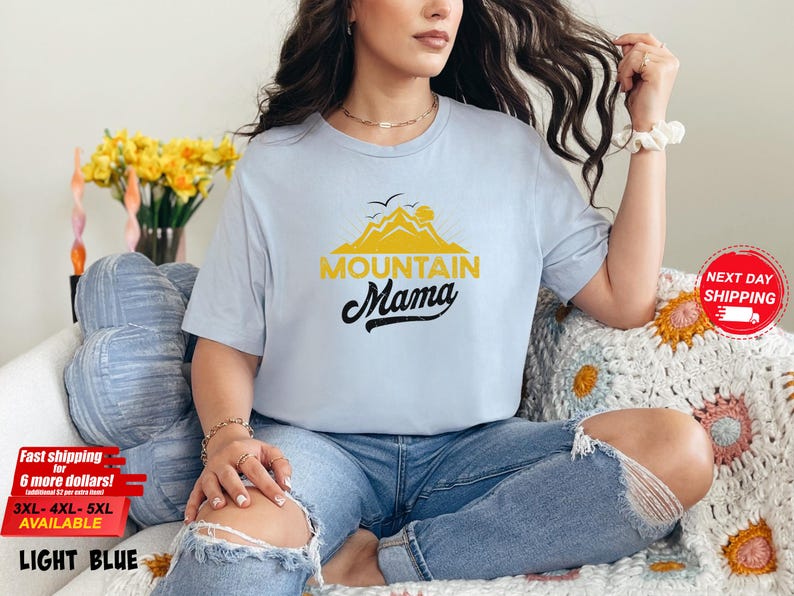 May include: Light blue t-shirt with the text "Mountain Mama" in black script and a yellow mountain graphic. The shirt is light blue and is available in sizes 3XL, 4XL, and 5XL.