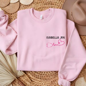 May include: Soft pink sweatshirt with the text "ISABELLA, RN" and a pink heart with a stethoscope graphic.