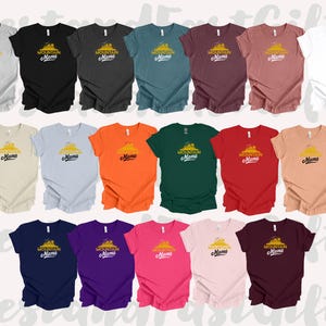May include: A collection of short-sleeved t-shirts in various colors, each featuring the text "Mountain Mama" with a mountain graphic in yellow and black. Colors include gray, black, teal, burgundy, and white.