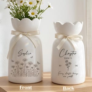 May include: Two white ceramic vases with a scalloped top and cream-colored ribbon. One vase has the name "Sophia" and a daisy illustration. The other vase says "Chapter Thirty" with a floral design and the text "Every chapter brings new growth."