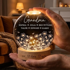 May include: A glowing glass sphere with the word "Grandma" and names of loved ones. The sphere sits on a wooden base with the words "we love you". The sphere has a floral design and is held in someone's hands.