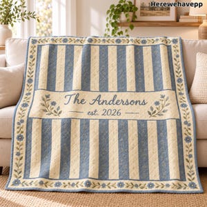 May include: A quilted throw blanket featuring alternating vertical stripes of cream and blue with a floral pattern. A central horizontal band displays the text The Andersons est. 2026 with floral accents. The border has a blue floral vine design.