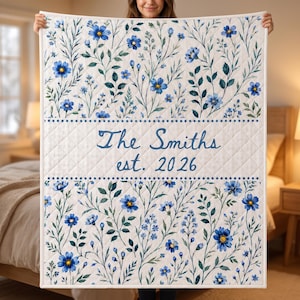 May include: A quilted throw blanket featuring a white background with a blue watercolor floral pattern. The center horizontal band contains the text The Smiths est. 2026 in blue script lettering, bordered by rows of small blue dots.