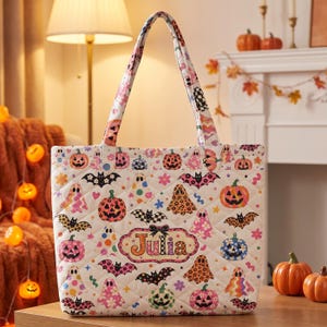 May include: A quilted tote bag featuring a white background with a colorful Halloween pattern of pumpkins, ghosts, bats, and flowers. The center of the bag displays a decorative label with the name Julia printed in bold orange letters.