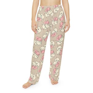Op de afbeelding: Beige pyjamabroek met een herhaald patroon van witte en roze cartoonhonden en harten. De broek is gemaakt van een zachte stof en heeft een losse pasvorm. De honden worden afgebeeld terwijl ze elkaar omhelzen.