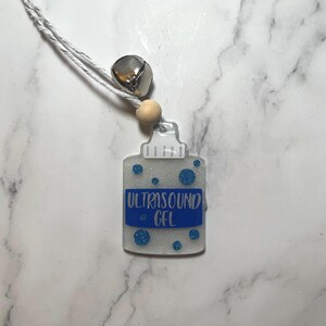 May include: A white and blue ultrasound gel bottle charm with a silver bell and a wooden bead on a white string.